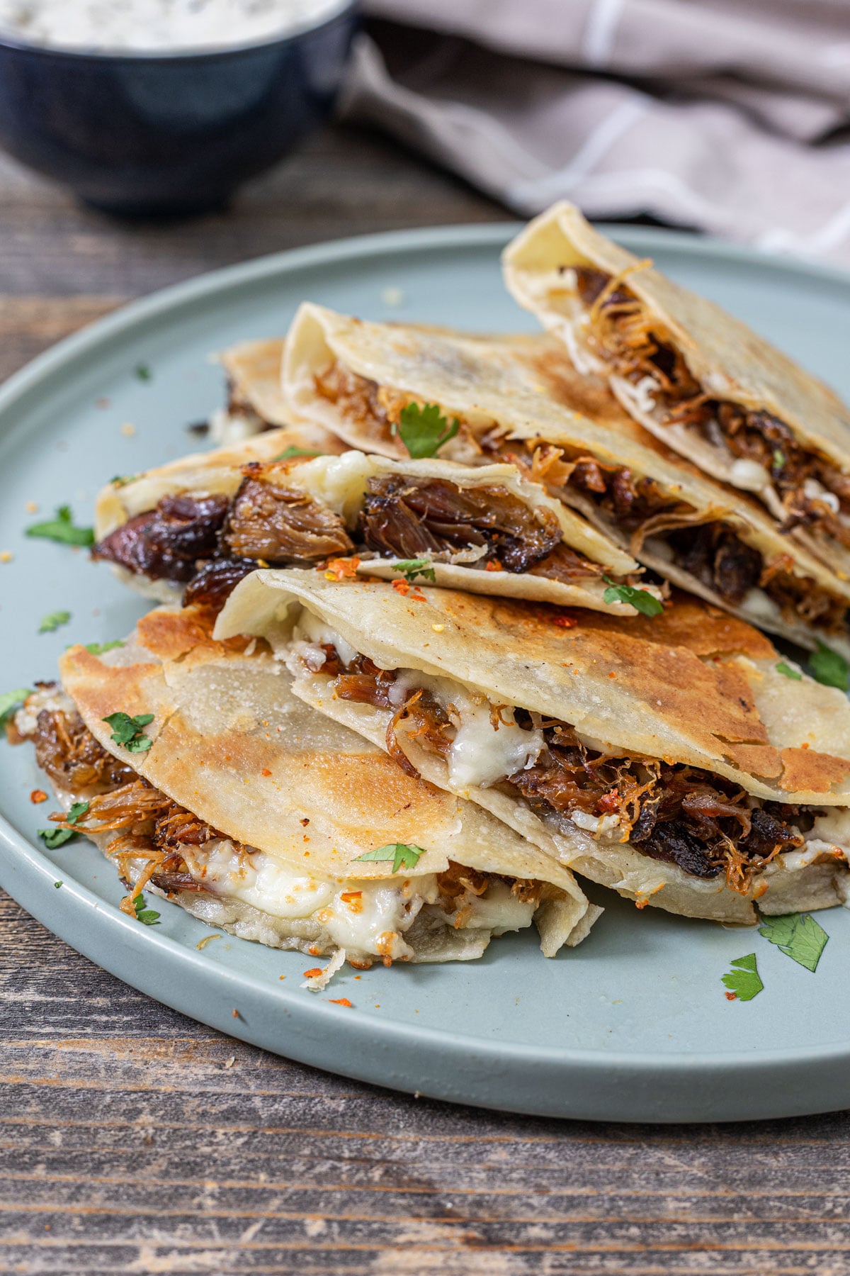 Smoked carnitas quesadilla wedges stacked on a plate with Oaxacan cheese visible in the layers and salsa verde crema in the background
