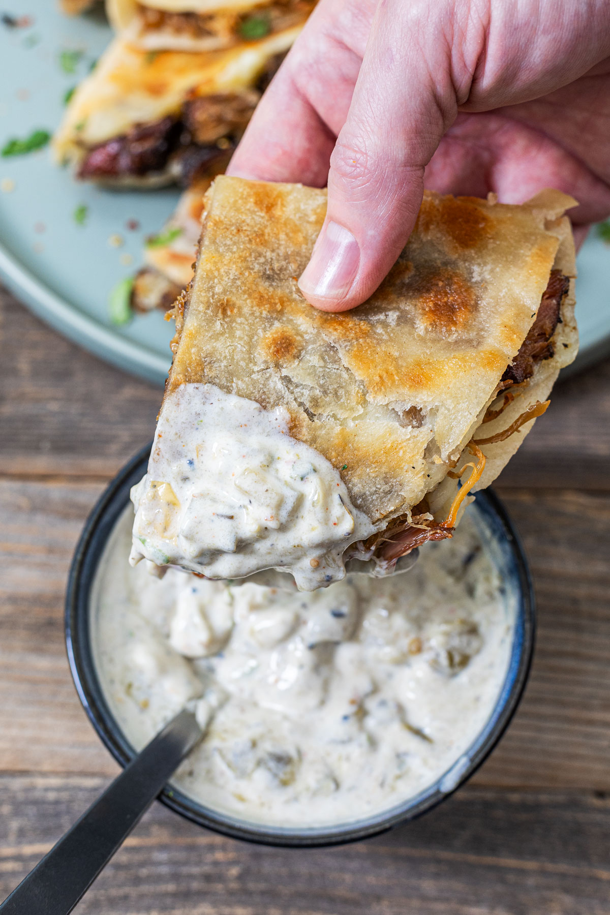 Hand dipping a smoked carnitas quesadilla wedge into a bowl of charred salsa verde crema