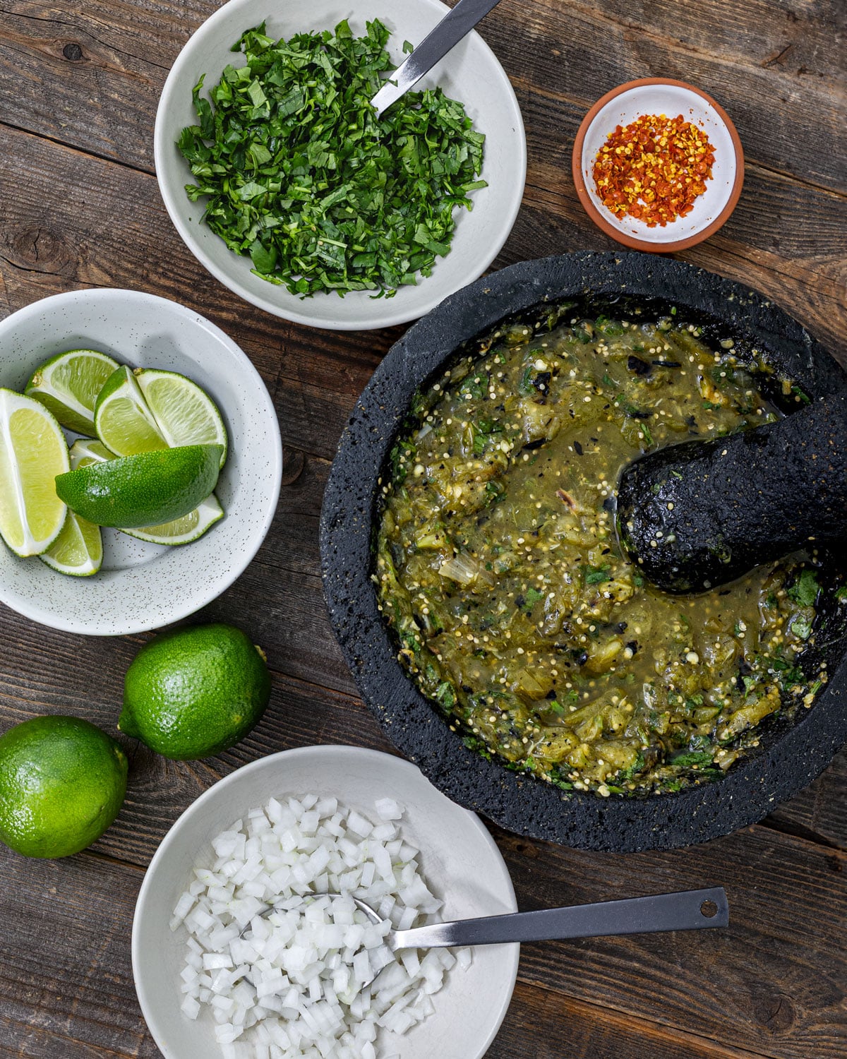 Carnitas taco toppings including charred salsa verde in a molcajete, diced white onion, cilantro, limes, and ground chiltepin