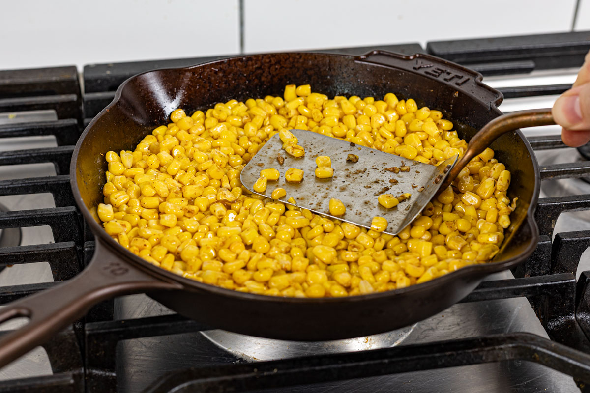 Corn kernels pressed flat in a cast iron skillet with a spatula, edges charring over high heat