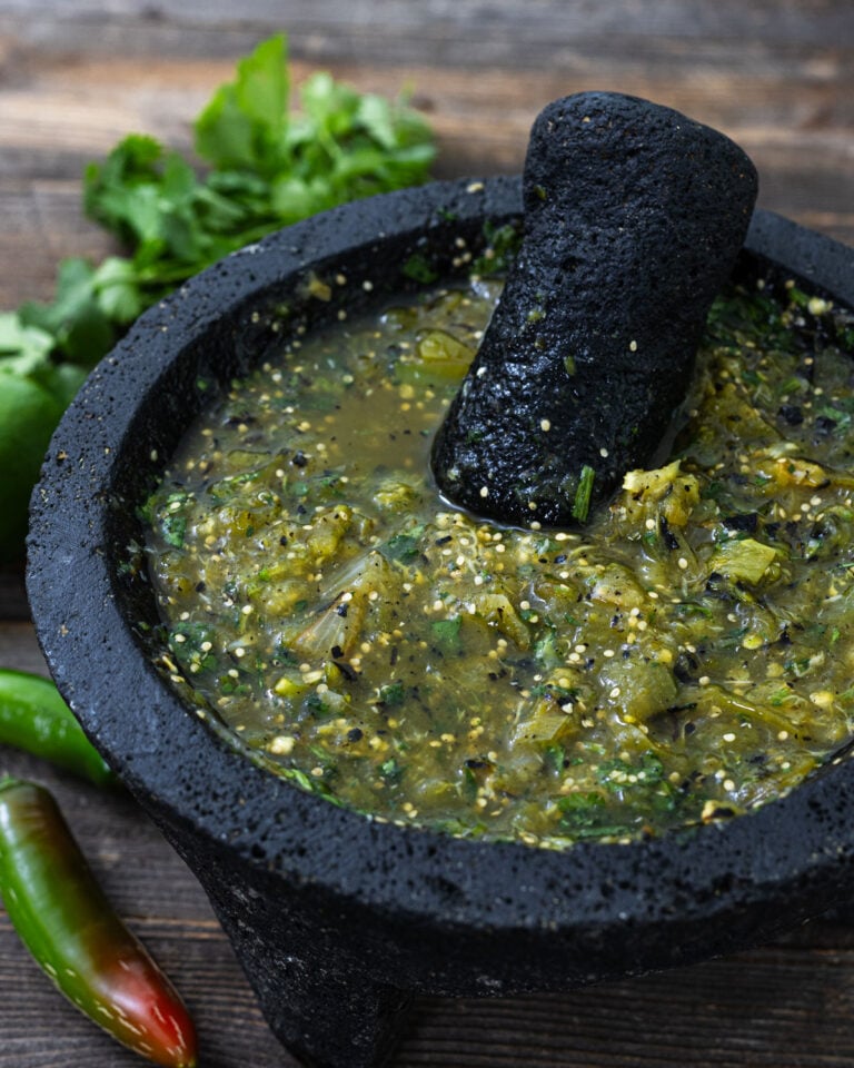 Charred salsa verde ground in a volcanic stone molcajete with tejolote, fresh serrano and jalapeno chiles, and cilantro on weathered wood