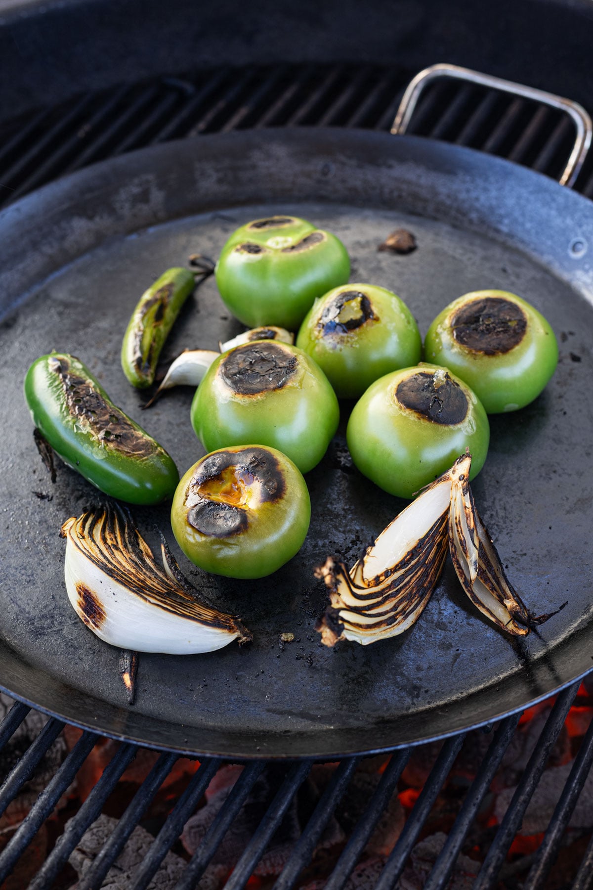 Tomatillos, serrano chile, and white onion charring on a carbon steel comal over live charcoal grill for charred salsa verde