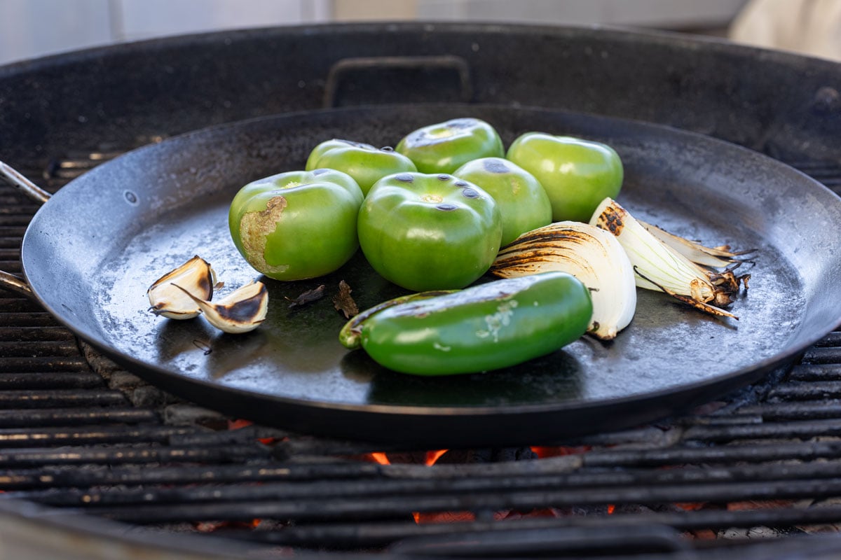 Tomatillos, jalapeño, white onion, and garlic on a carbon steel comal over live charcoal at the start of charring for salsa verde,