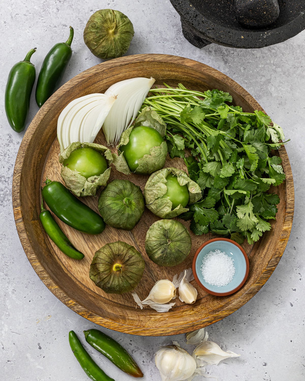 Fresh salsa verde ingredients on a wood plate including tomatillos with husks, serrano and jalapeno chiles, white onion, cilantro, garlic, and salt