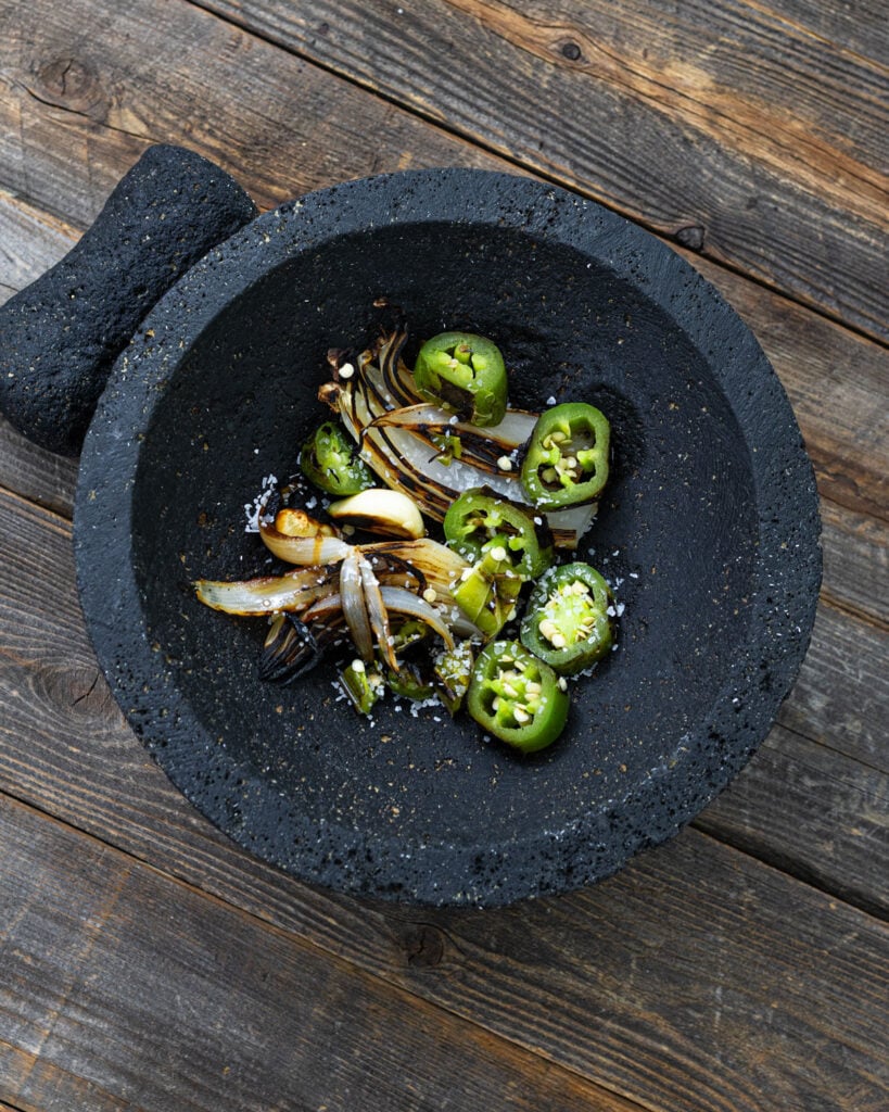 Charred serrano chiles, roasted onion, and peeled garlic with salt in a volcanic stone molcajete before grinding