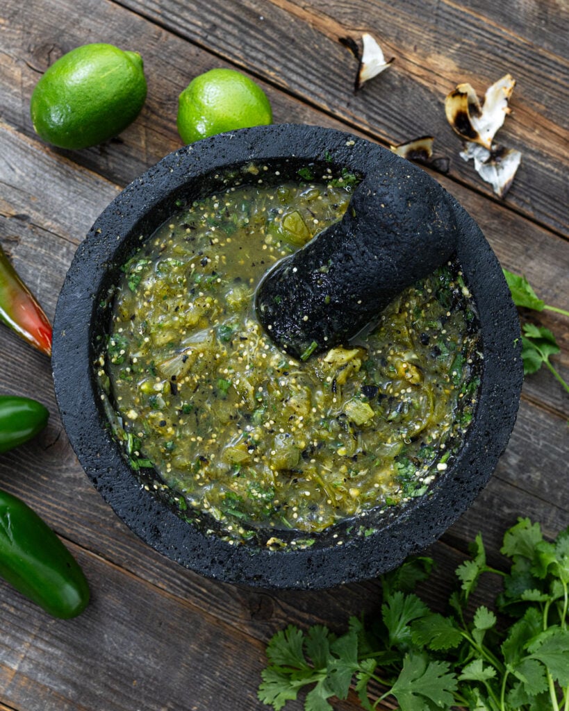 Charred salsa verde freshly ground in a volcanic stone molcajete with tejolote, charred garlic skins, limes, and cilantro on weathered wood
