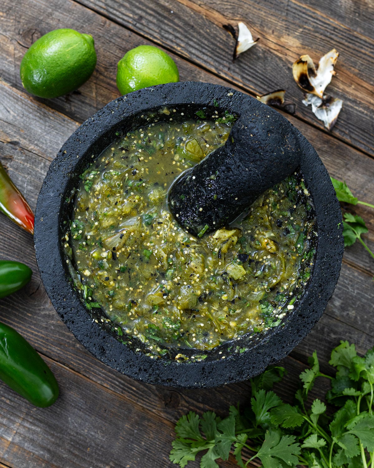 Charred salsa verde freshly ground in a volcanic stone molcajete with tejolote, charred garlic skins, limes, and cilantro on weathered wood