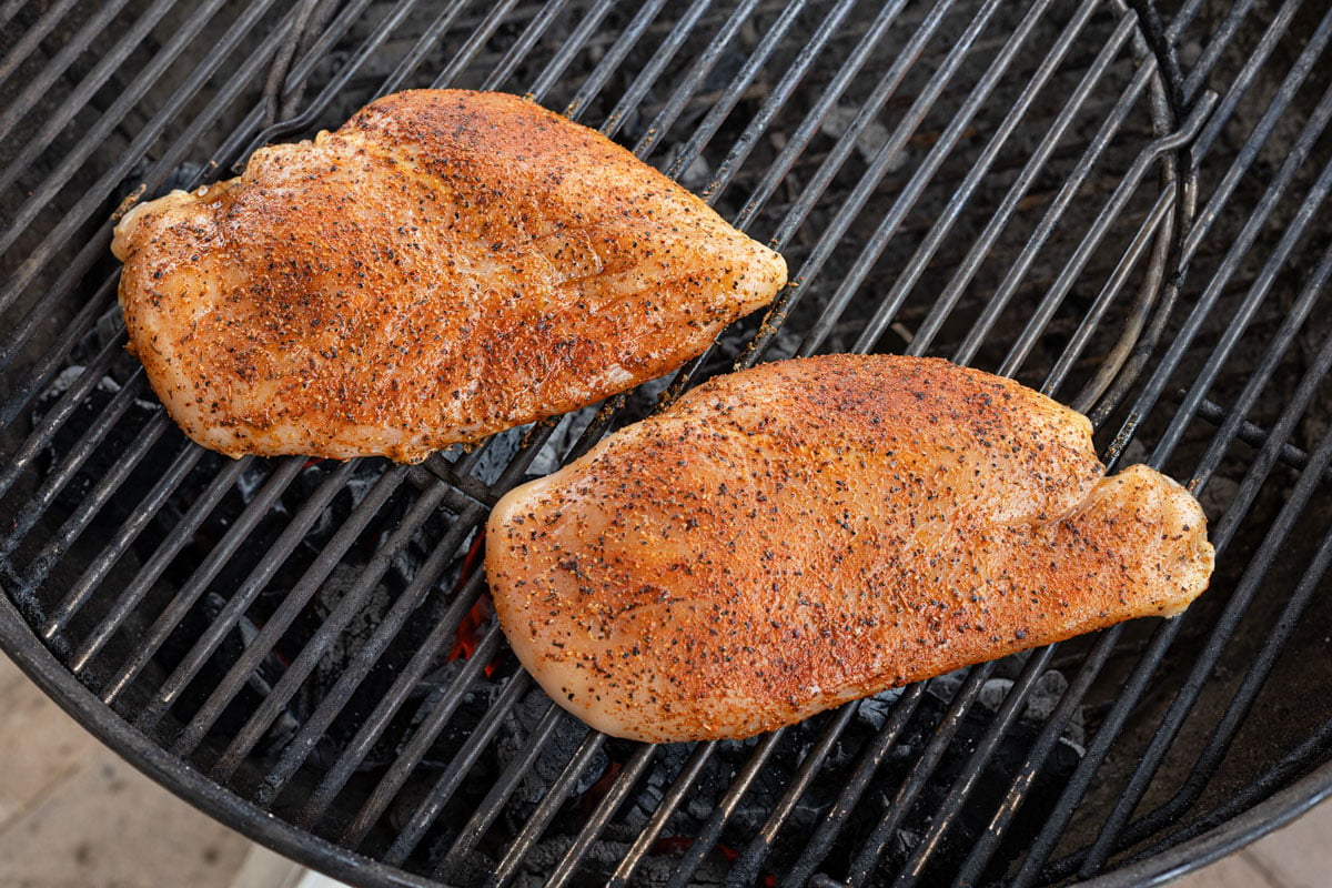Two seasoned chicken breasts finishing on the indirect side of a charcoal kettle grill