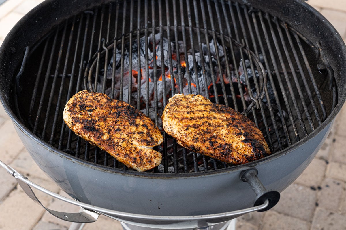 Two grilled chicken breasts resting on the indirect zone of a charcoal kettle grill with glowing coals visible on the direct side