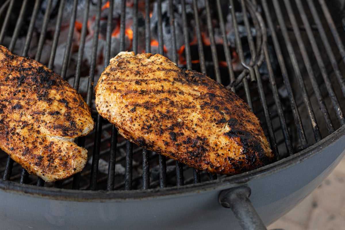 Two chicken breasts with grill marks moving to the indirect side of a charcoal kettle grill to finish cooking