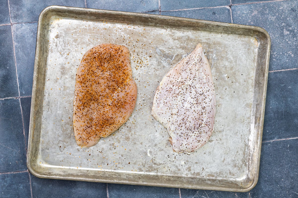 Two chicken breasts on a sheet pan showing the oil and seasoning method on the left versus the mayonnaise method on the right