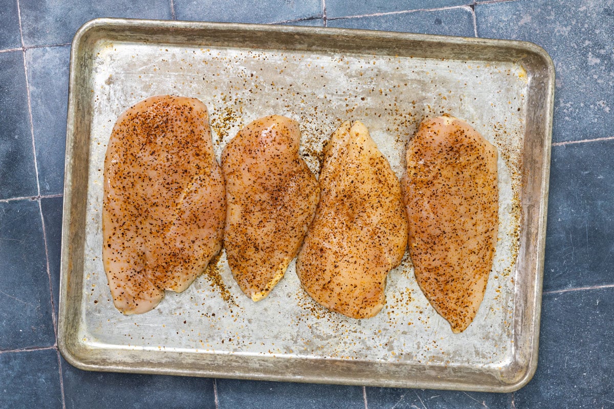 Four seasoned chicken breasts on a sheet pan ready for the grill