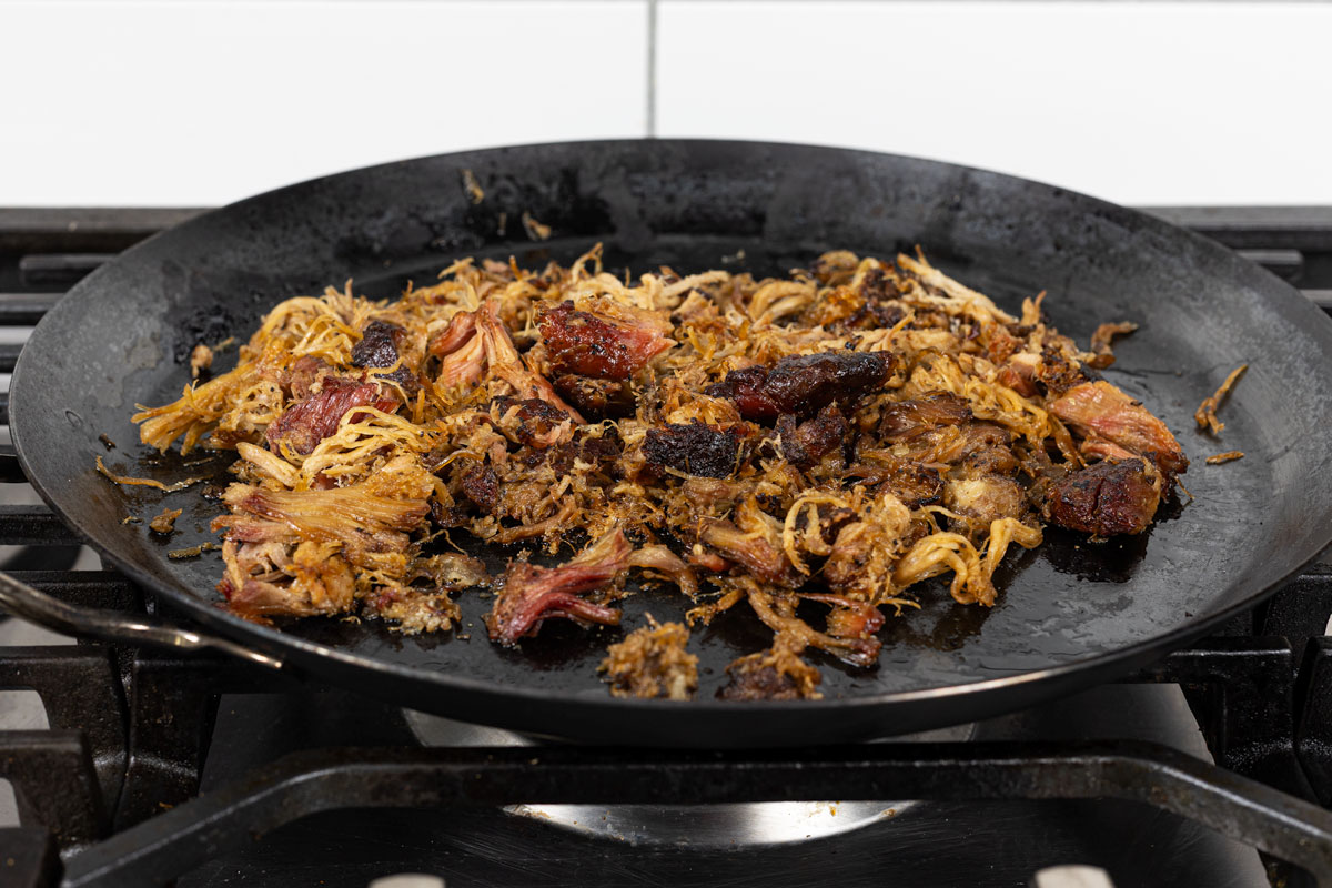 Smoked carnitas crisping on a large black comal over a gas burner, edges caramelized
