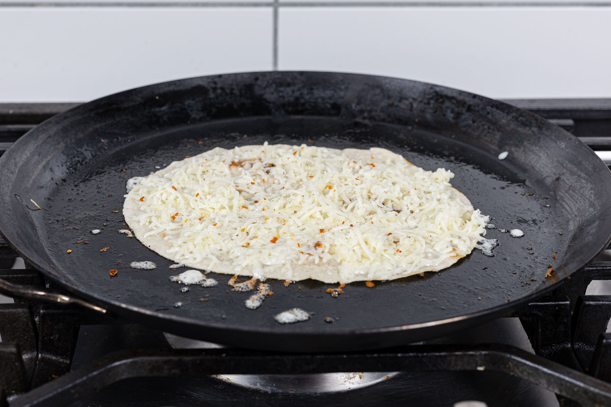 Oaxacan cheese spread across a flour tortilla on a black comal over a gas burner