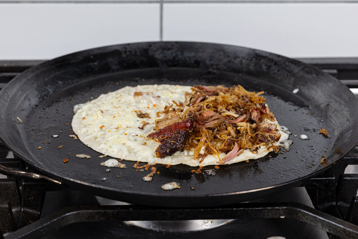 Smoked carnitas added to one half of an Oaxacan cheese tortilla on a comal, ready to fold