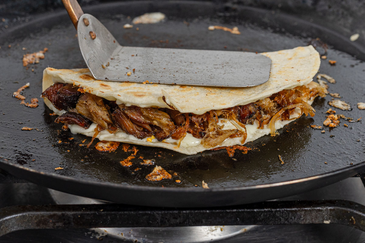 Smoked carnitas quesadilla folded and pressed with a spatula on a black comal, cheese melting at the edges
