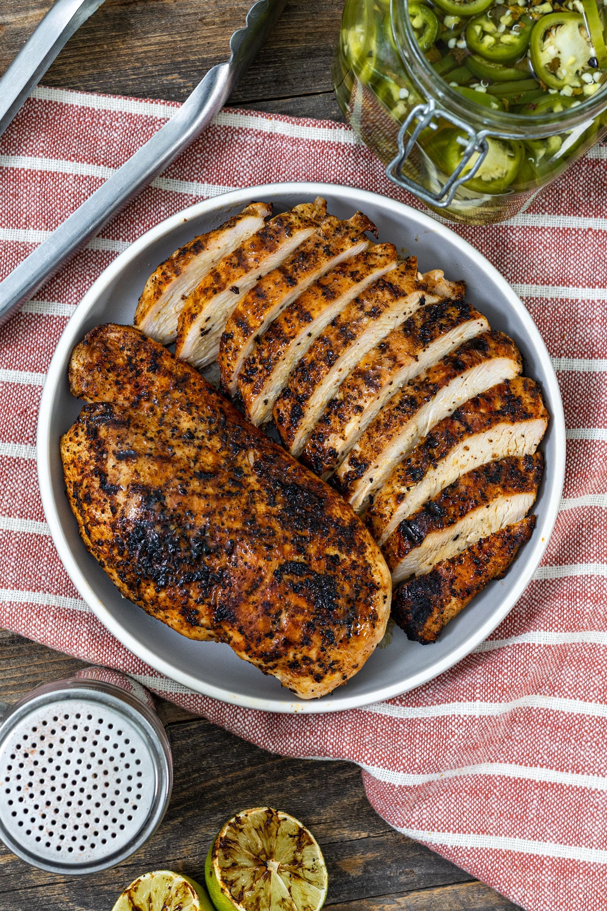 Grilled chicken breast sliced on a plate with pickled jalapeño jar, grilled limes, and seasoning shaker on a wood panel surface