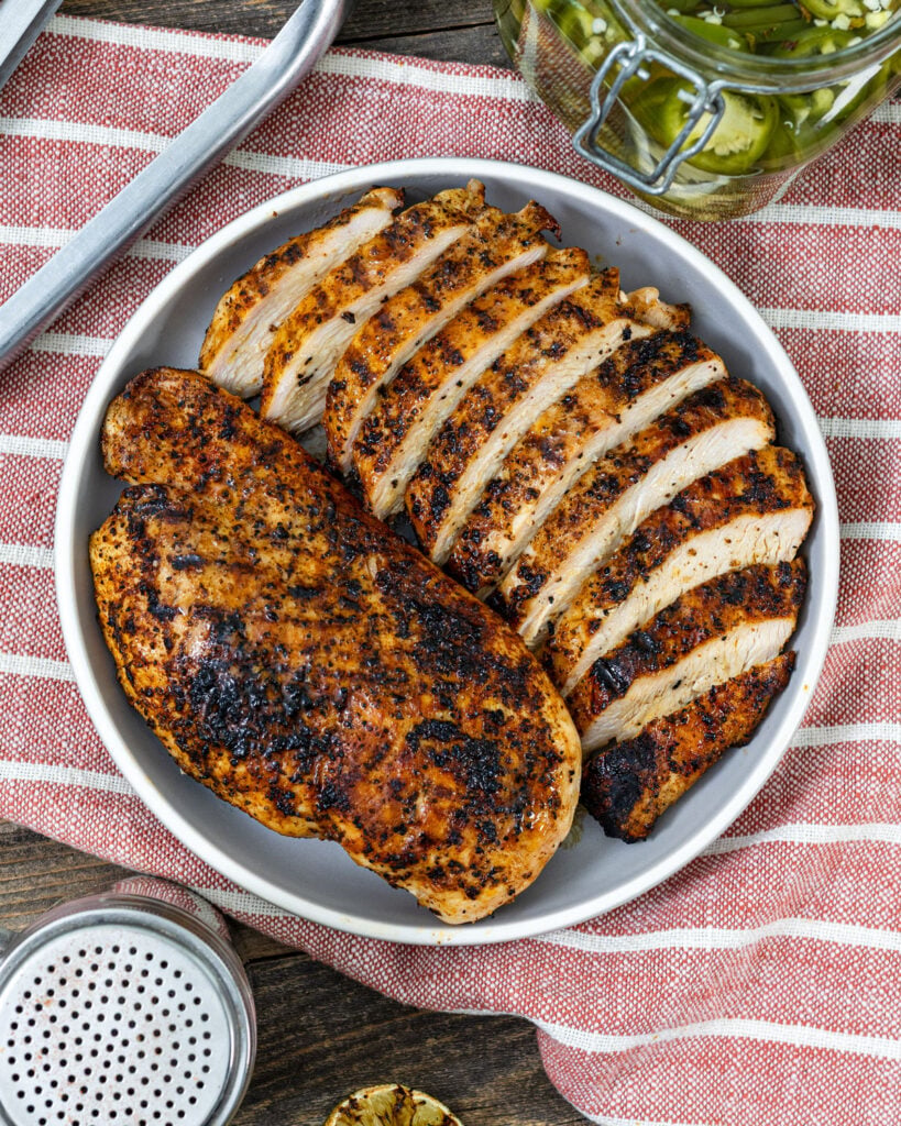 Grilled chicken breast sliced on a plate with pickled jalapeño jar, grilled limes, and seasoning shaker on a wood panel surface