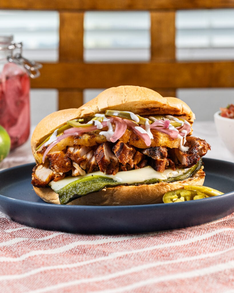 Grilled chicken torta on a blue plate with cheese-stuffed poblano, pickled red onions, jalapeños, and cilantro lime crema on a bolillo roll