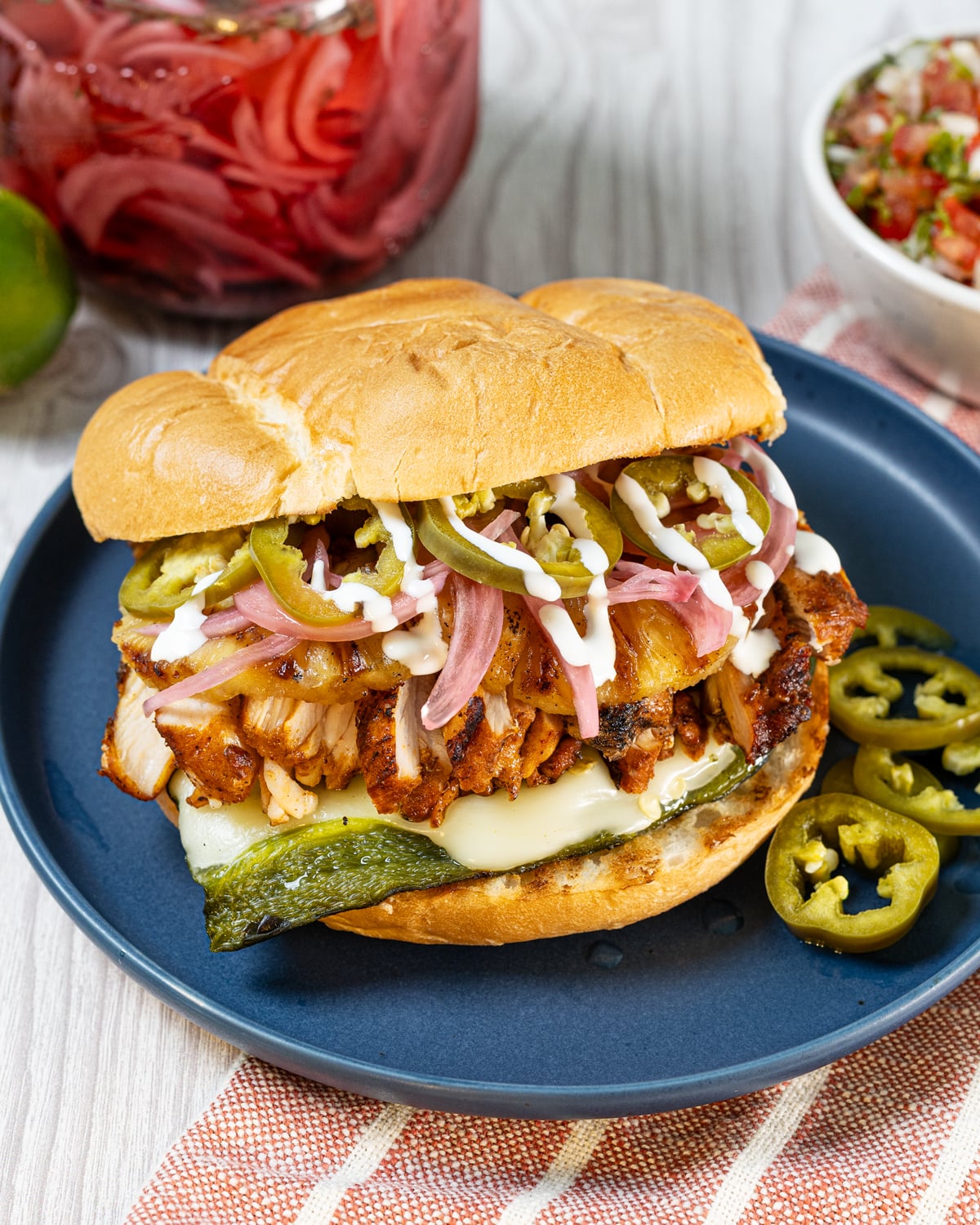 Grilled chicken torta stacked on a blue plate with grilled pineapple, pickled jalapeños, pickled red onions, and cilantro lime crema