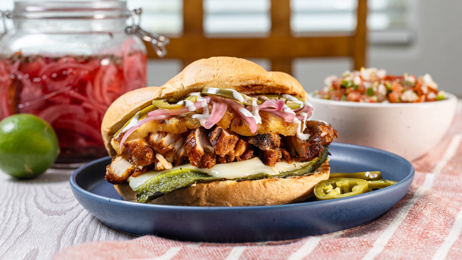 Grilled chicken torta on a blue plate with stuffed poblano and pickled toppings, served with pico de gallo and pickled red onions in the background