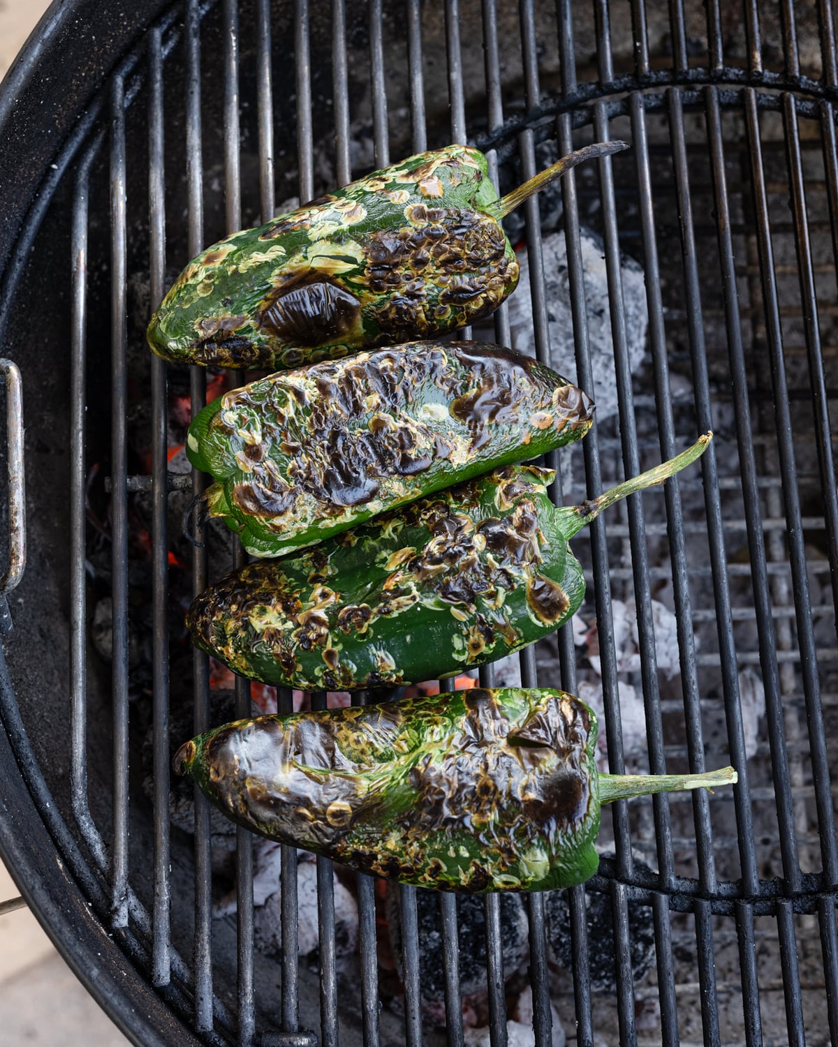 Whole poblano peppers charring directly over live charcoal on a kettle grill
