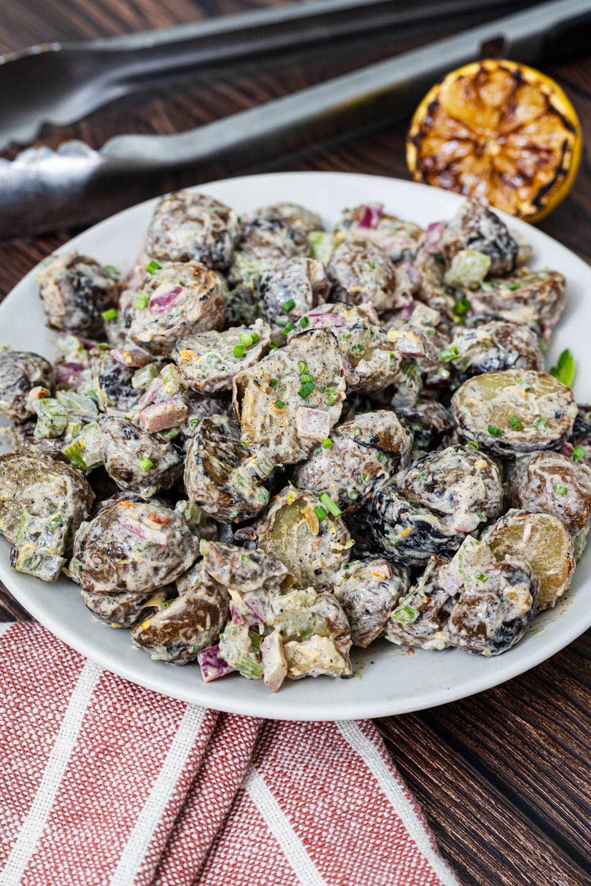 Grilled potato salad on a white plate with charred mini potatoes, grilled red onion, celery, lemon zest, and fresh chives, grilled lemon and tongs in background