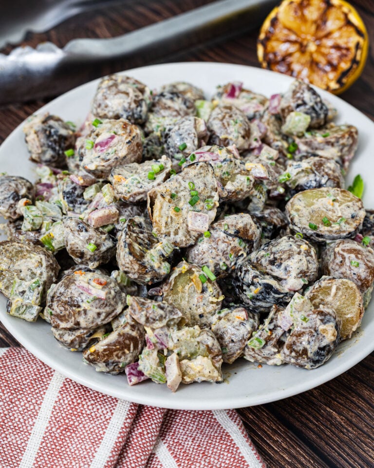 Grilled potato salad on a white plate with charred mini potatoes, grilled red onion, celery, lemon zest, and fresh chives, grilled lemon and tongs in background