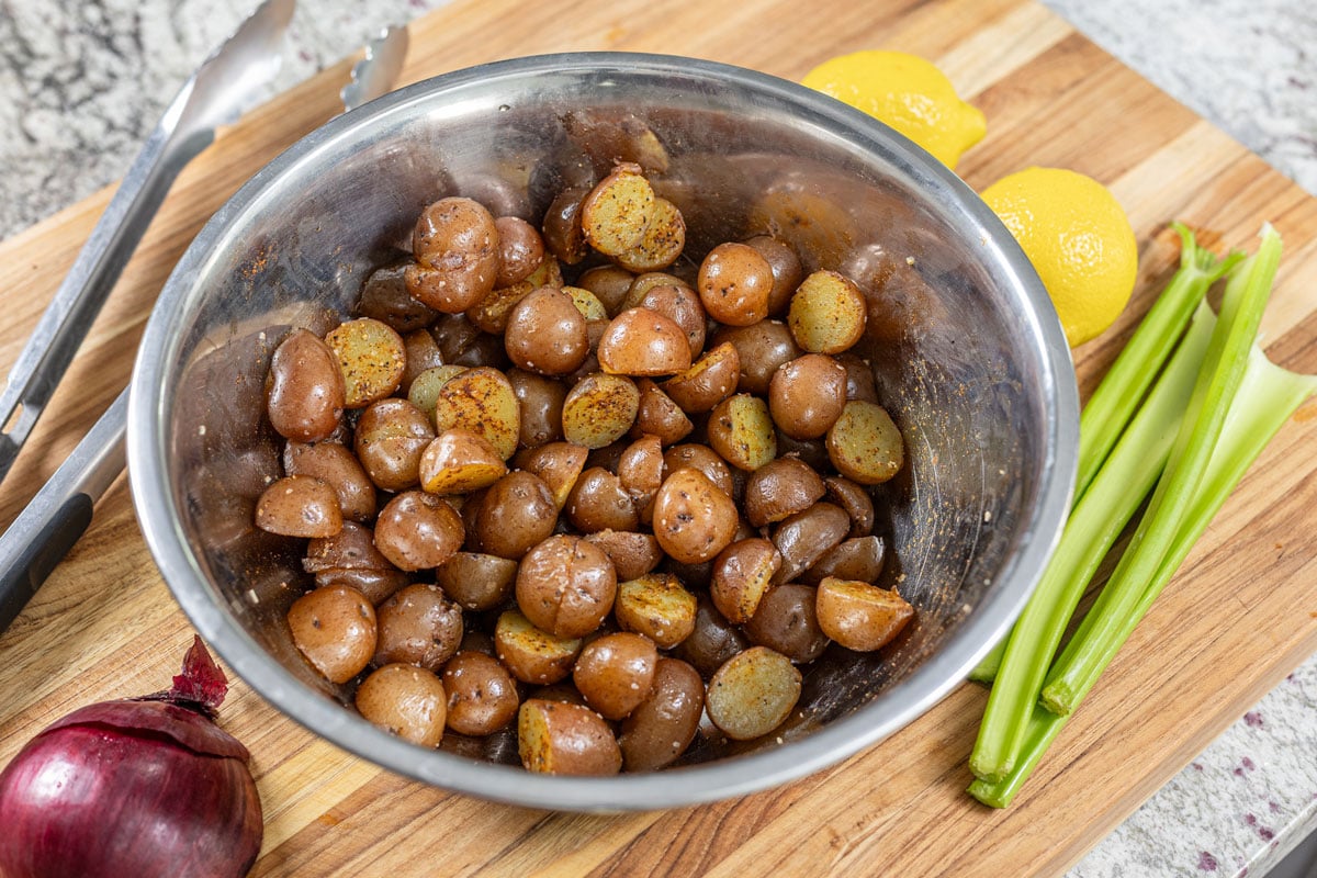 Halved mini potatoes tossed in olive oil, paprika, and black pepper in a metal bowl with celery, lemons, and red onion alongside