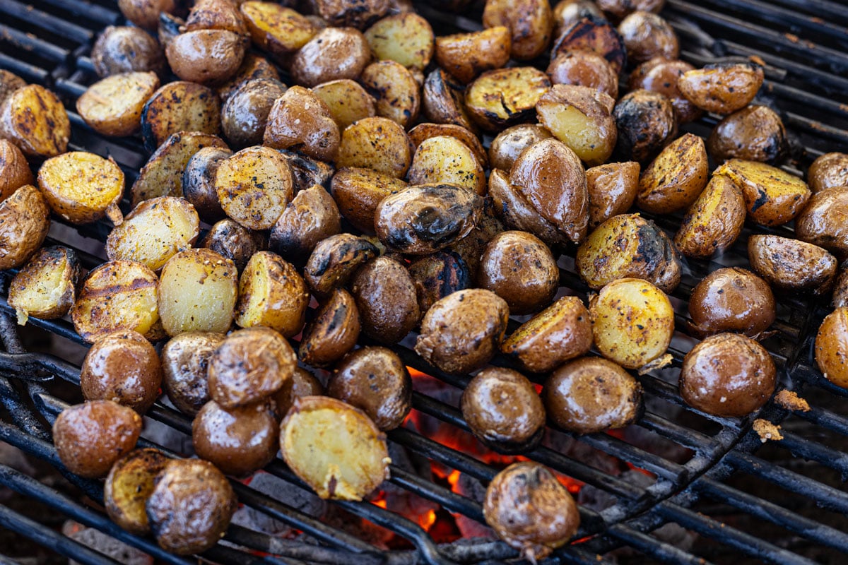 Halved mini potatoes cut-side down on charcoal grill grates with visible char and live fire below