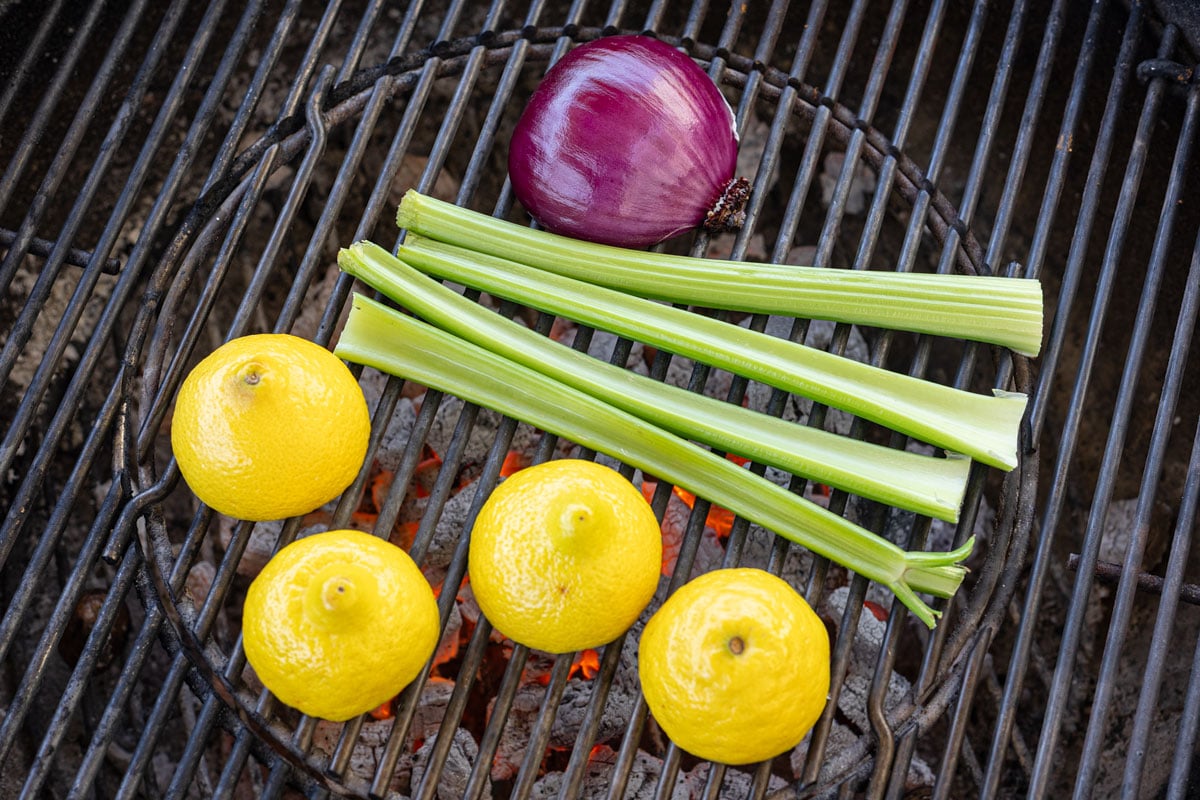 Whole lemons, celery stalks, and red onion half on charcoal grill grates before grilling