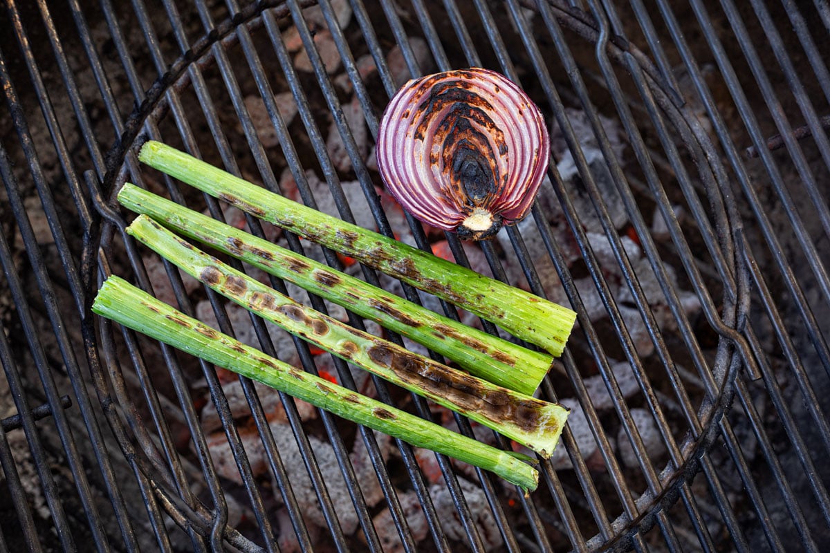 Grilled celery stalks and red onion half with char marks on charcoal grill grates