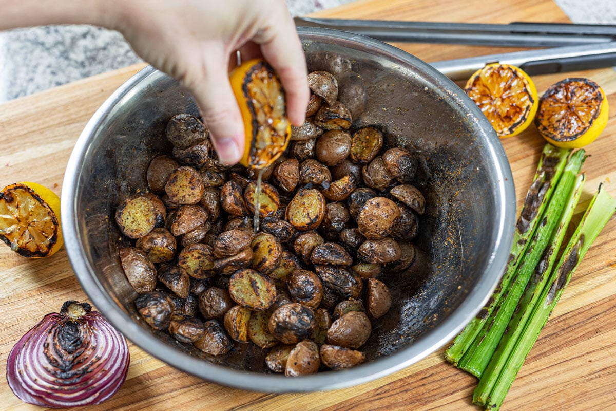 Hand squeezing a grilled lemon half over charred mini potatoes in a metal bowl, grilled vegetables alongside