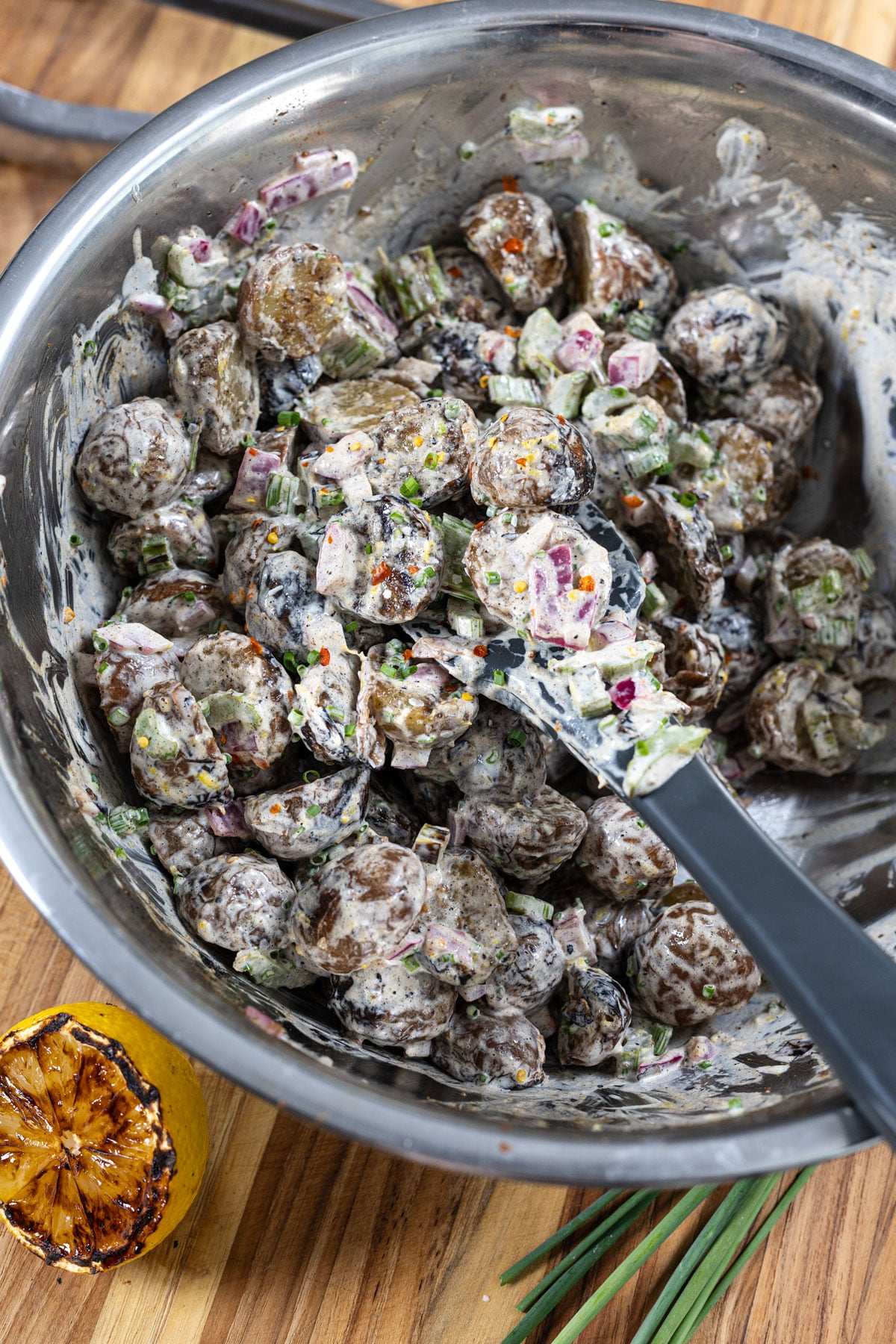 Grilled potato salad being folded together in a metal mixing bowl with dressing, chives, and grilled vegetables