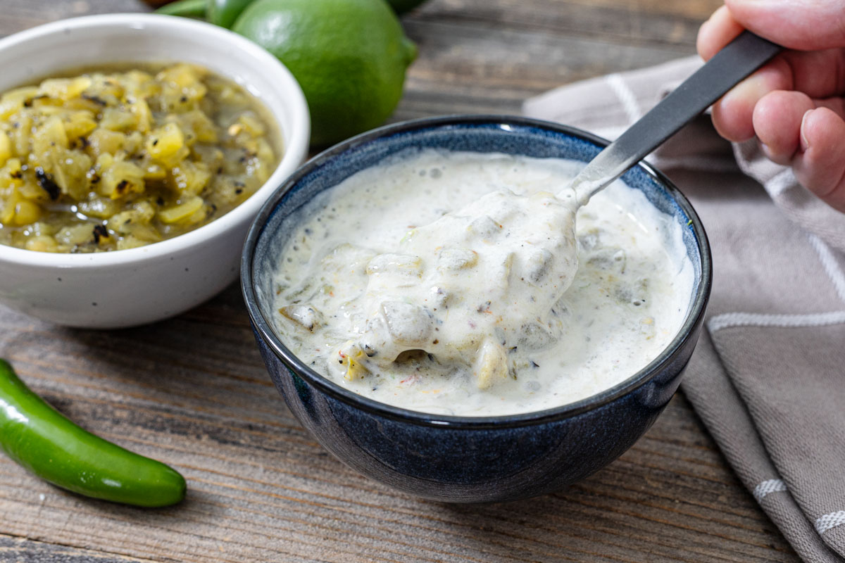 Salsa verde crema in a dark blue bowl with a spoon lifting the sauce, charred salsa verde and lime in the background