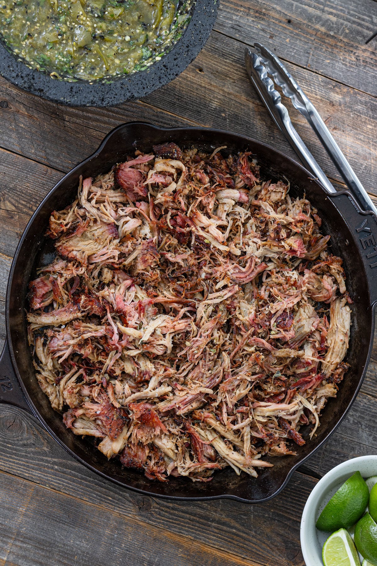 Shredded smoked carnitas in a cast iron skillet beginning to crisp with salsa verde and limes alongside