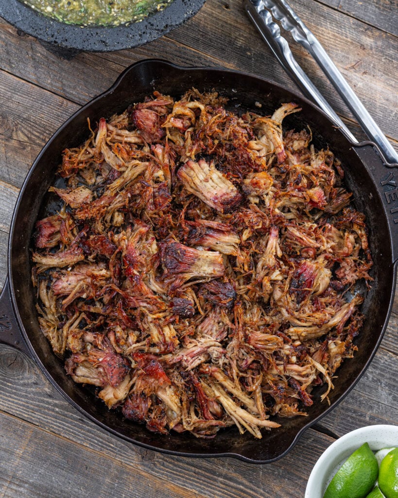 Shredded smoked carnitas crisping in a cast iron skillet with caramelized edges, salsa verde, and lime wedges