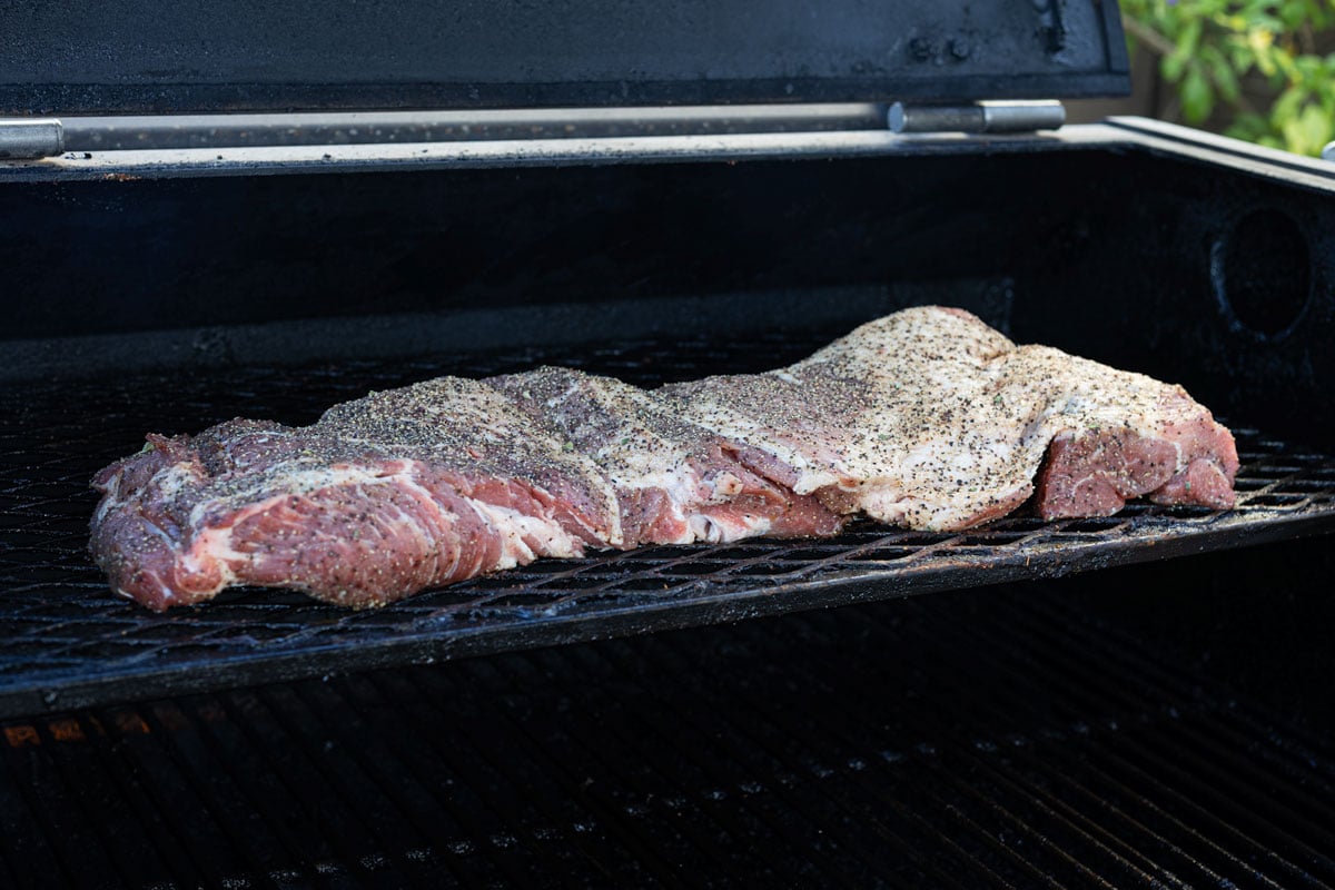 Butterflied pork shoulder seasoned with carnitas spice blend flat on smoker grates at the start of the cook