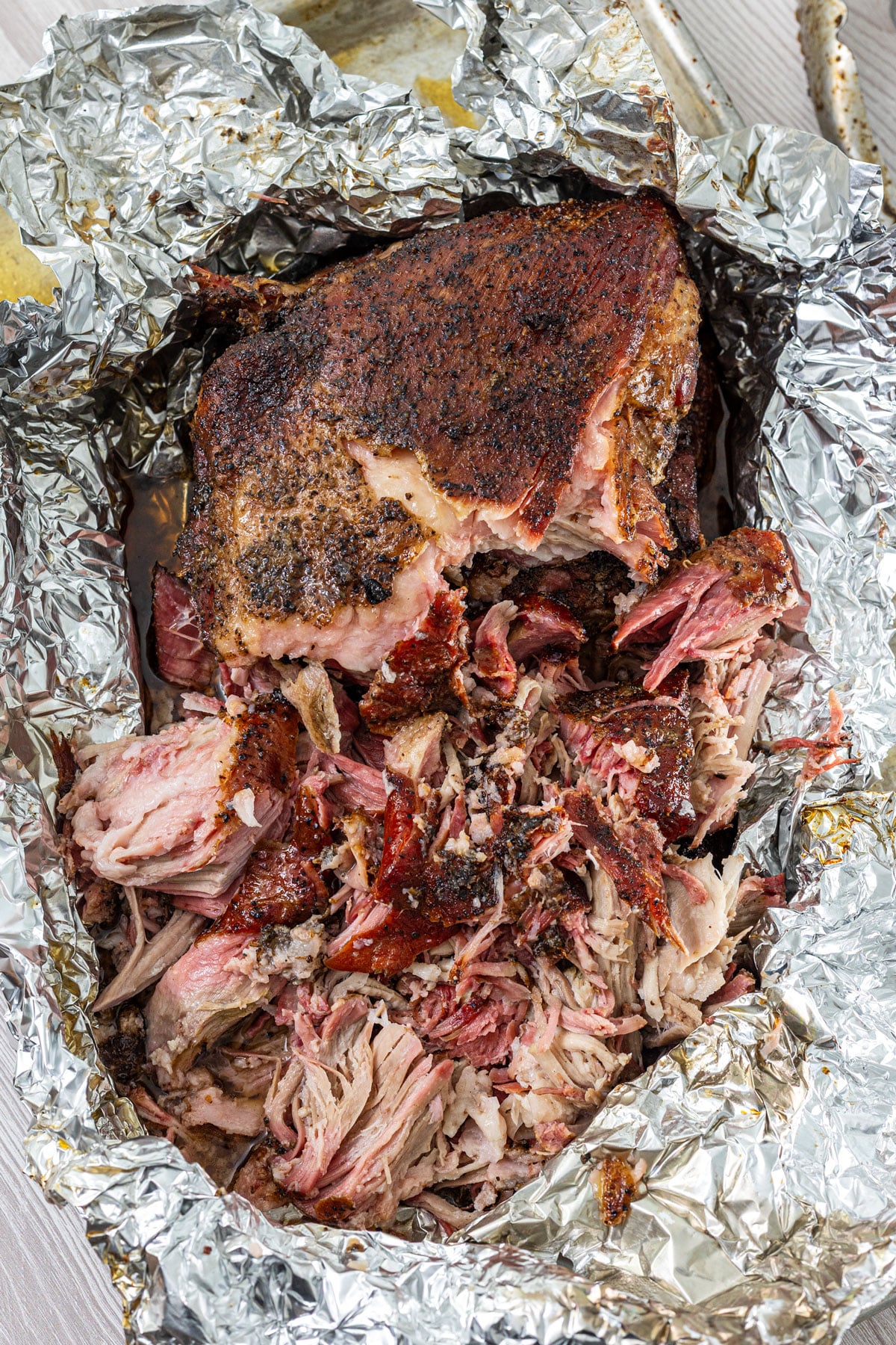 Smoked carnitas pork shoulder being shredded in foil with dark bark pieces throughout