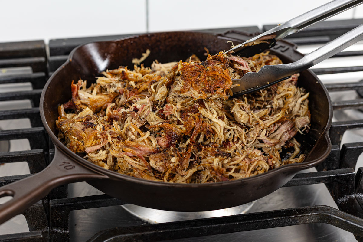 Smoked carnitas crisping in a cast iron skillet with tongs lifting caramelized shredded pork