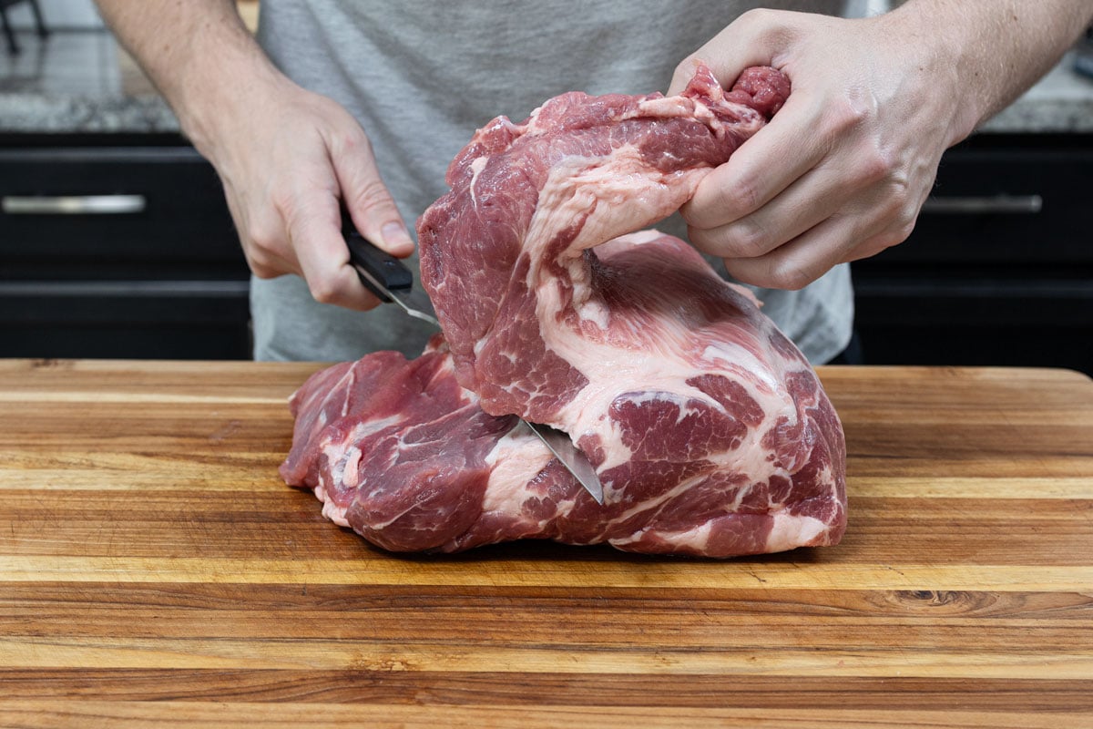 Pork shoulder being butterflied along the natural muscle seam with a boning knife on a wood cutting board