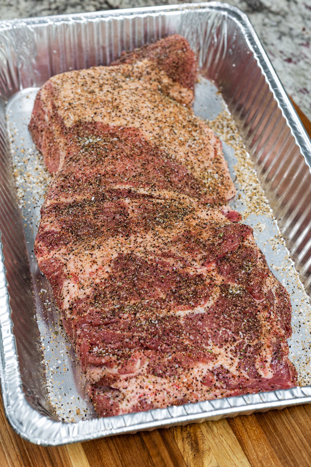 Butterflied pork shoulder coated in carnitas spice blend in an aluminum pan ready for the smoker