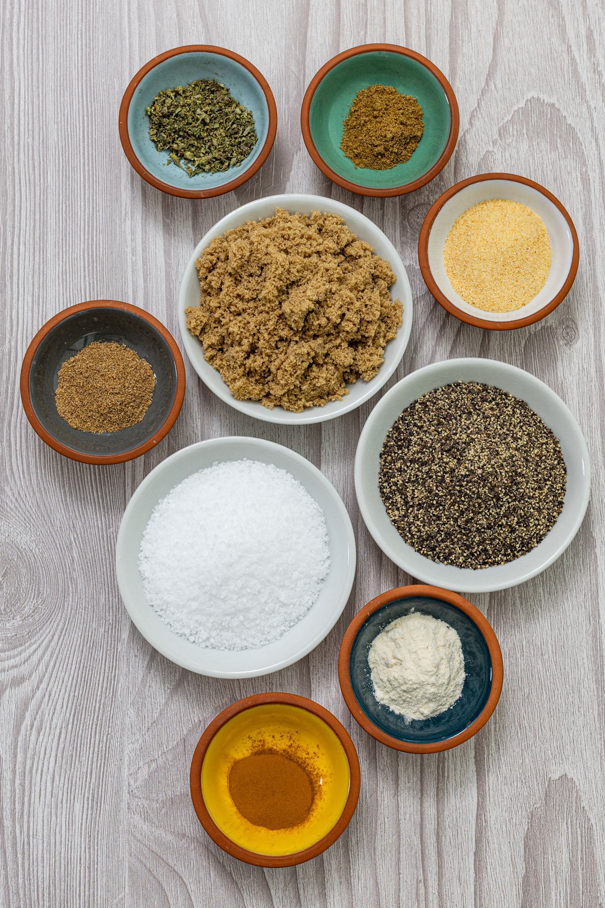 Carnitas spice blend ingredients in individual bowls including brown sugar, 16-mesh black pepper, kosher salt, Mexican oregano, cumin, coriander, granulated garlic, onion powder, and cinnamon
