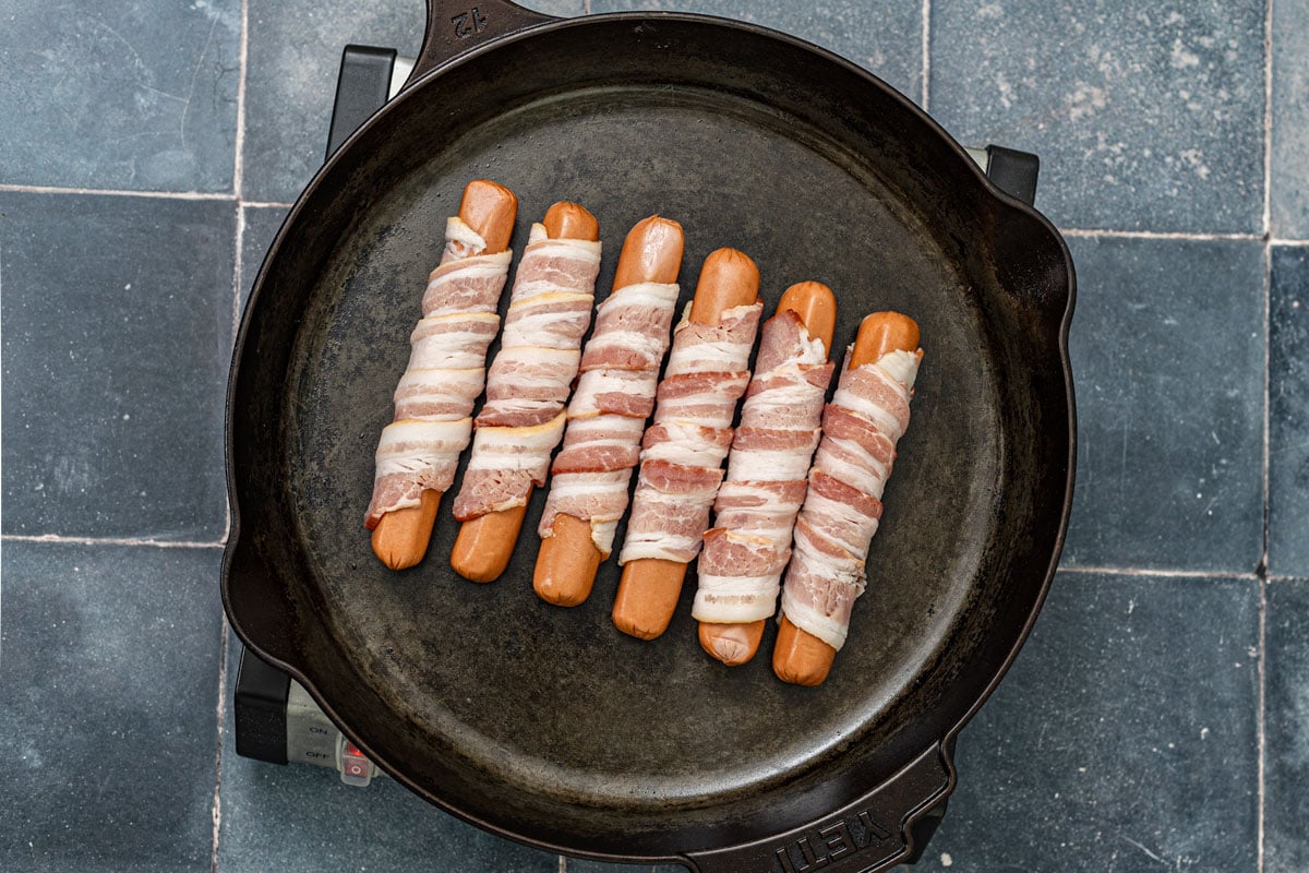 Bacon-wrapped hot dogs placed seam-side down in a cast iron skillet before cooking