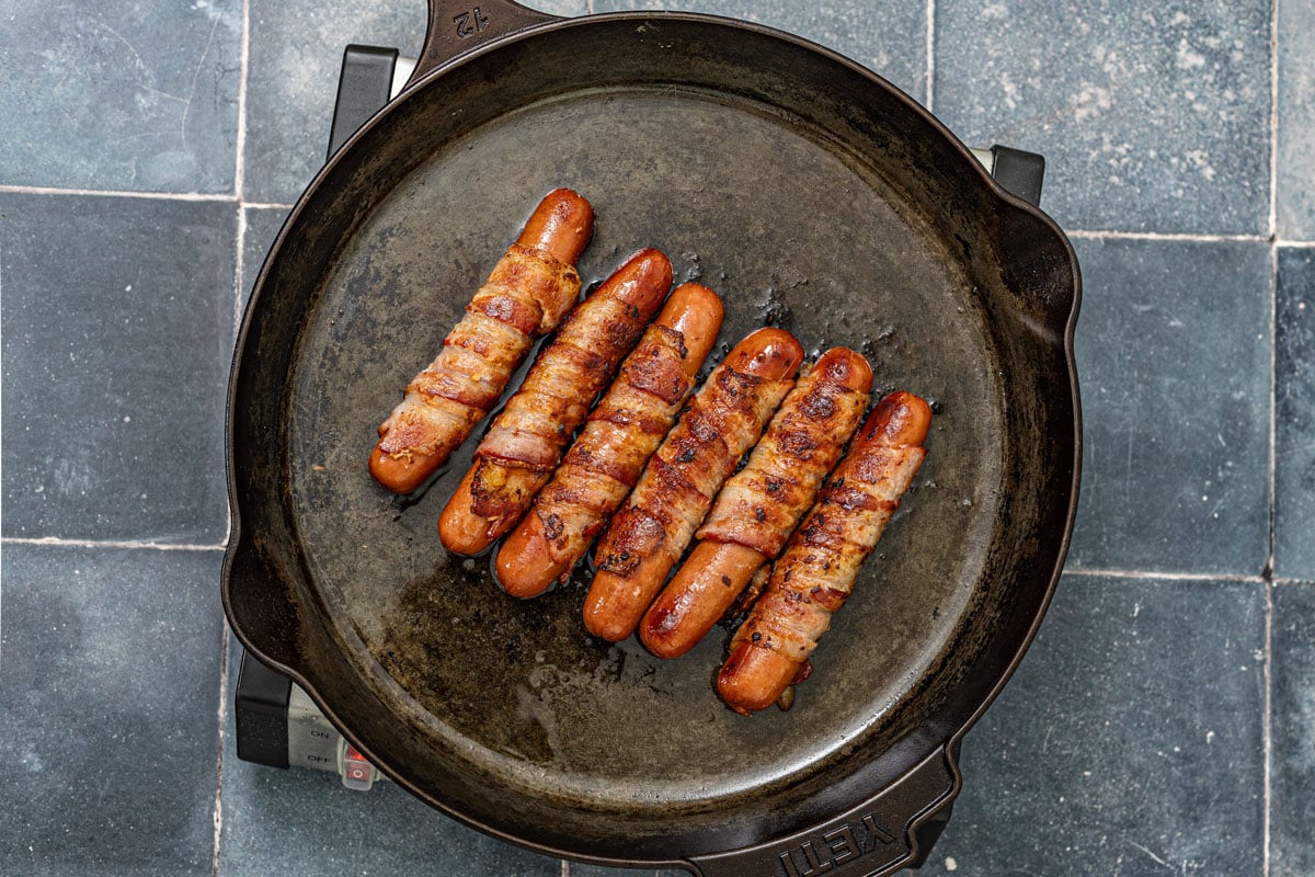 Bacon-wrapped hot dogs cooked until crispy in a cast iron skillet over medium heat