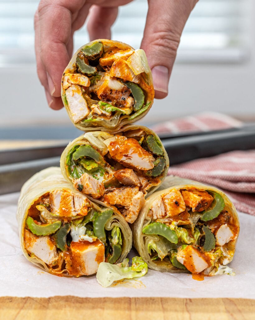 Hand picking up a buffalo chicken wrap from a stack showing grilled chicken, buffalo sauce, and grilled celery filling