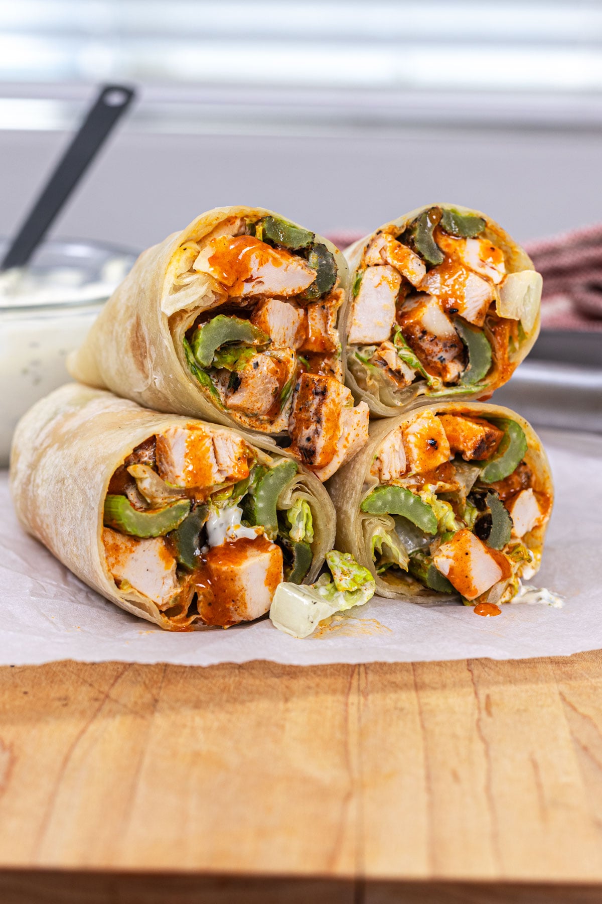 Buffalo chicken wraps sliced and stacked on parchment showing grilled chicken, buffalo sauce, and grilled celery filling with blue cheese dressing in the background