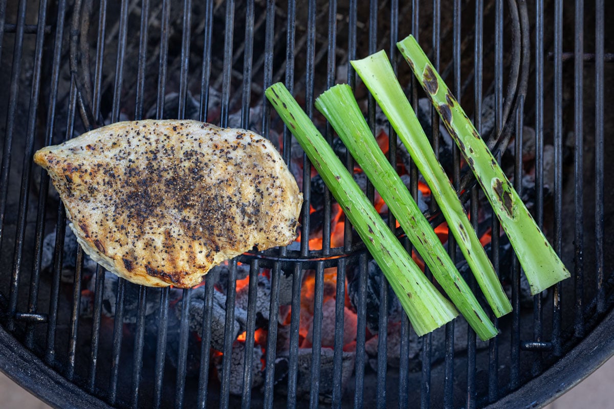 Chicken breast and celery stalks grilling side by side over live charcoal on a kettle grill