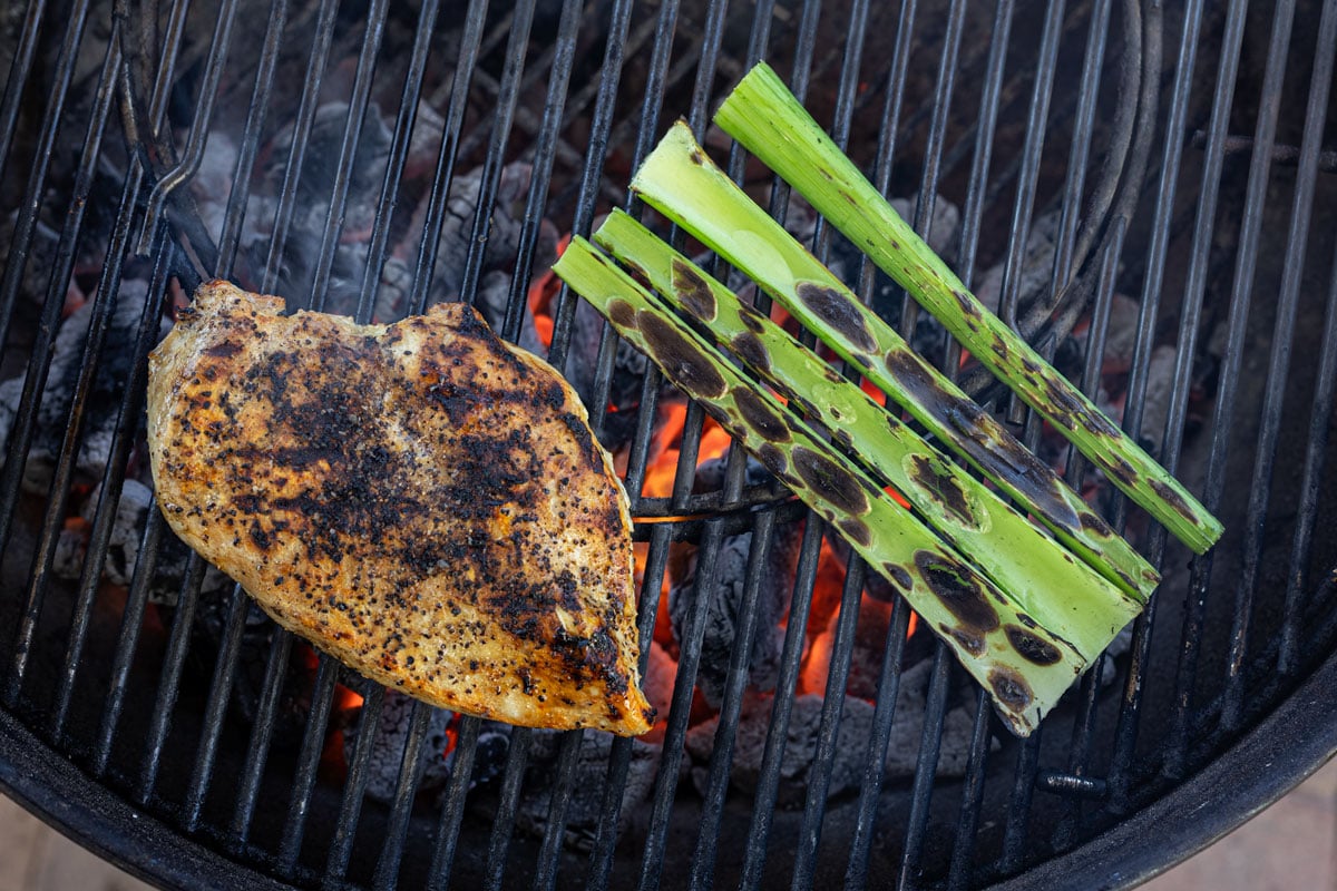 Celery stalks developing char marks over live charcoal on a kettle grill alongside a grilled chicken breast
