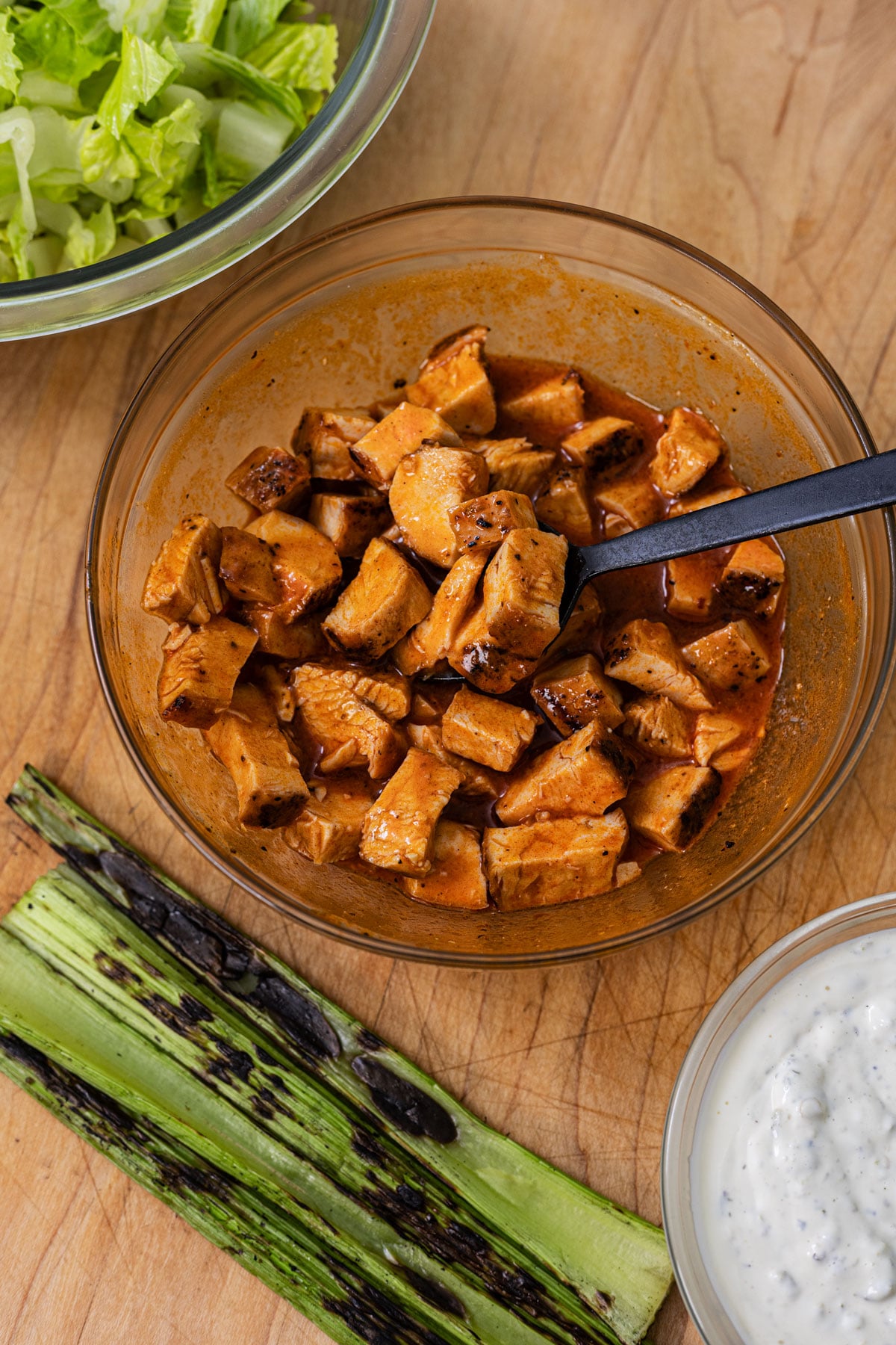 Cubed grilled chicken tossed in buffalo sauce in a glass bowl with grilled celery stalks and blue cheese dressing on a cutting board